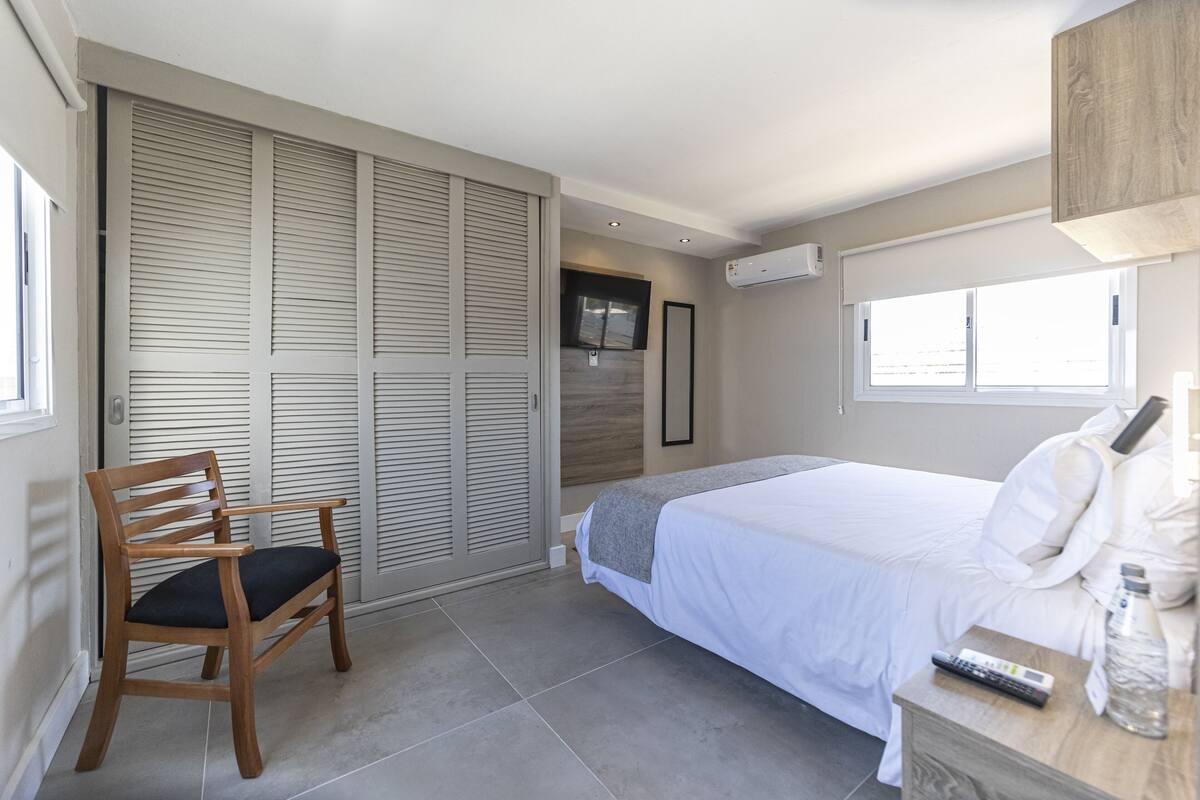 A bedroom with a bed, a wooden chair, a bedside table with a bottle, and a window.