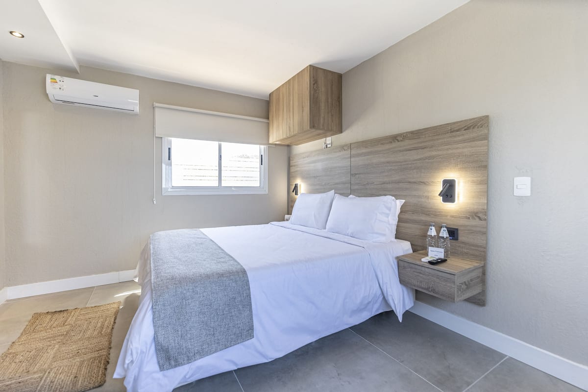 A modern bedroom with a bed, bedside table, wall-mounted light, and an air conditioning unit.