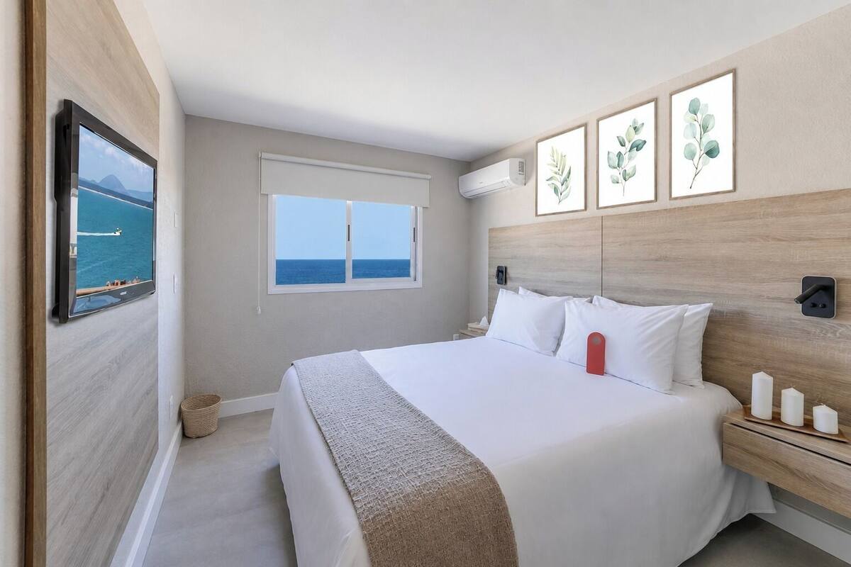 A hotel room with a bed, a TV, a window with a view of the sea, and wall-mounted artwork.