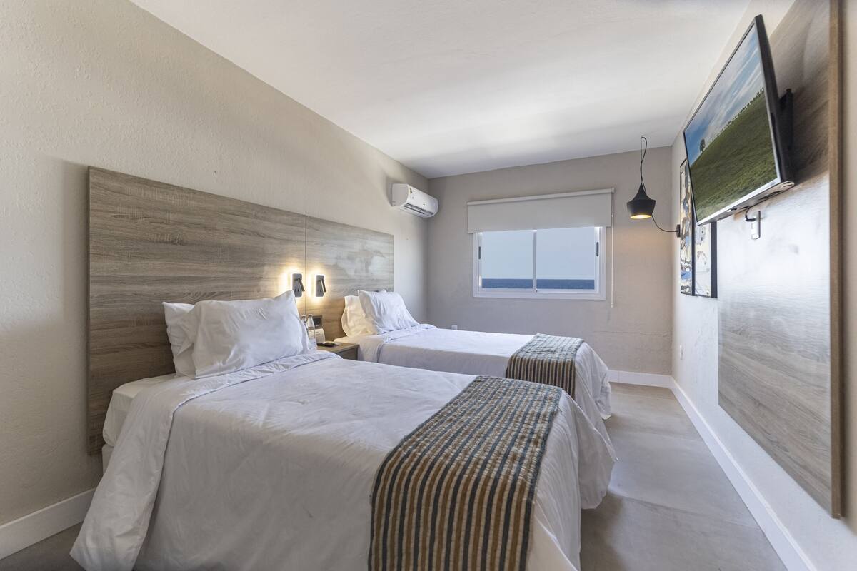 A hotel room with two beds, a large window, an air conditioning unit, and a flat-screen TV mounted on the wall.