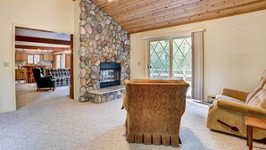 Un salon chaleureux avec une cheminée en pierre, un plafond en bois et une vue dégagée sur l’extérieur grâce à une grande fenêtre.
