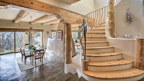 A spacious interior with a wooden staircase, a dining area with a table and chairs, and large windows offering a view of the outdoors. - Adobe Home - River & Mtn Views w/ Hot Tub! (Redmond)