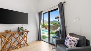 A modern living room with a flat-screen TV, wooden coffee table, and a grey sofa with floral cushions. A sliding glass door leads to a pool area with outdoor furniture. - Cove Oasis: Pool, Hikes, Golf, Festival Fun (La Quinta)