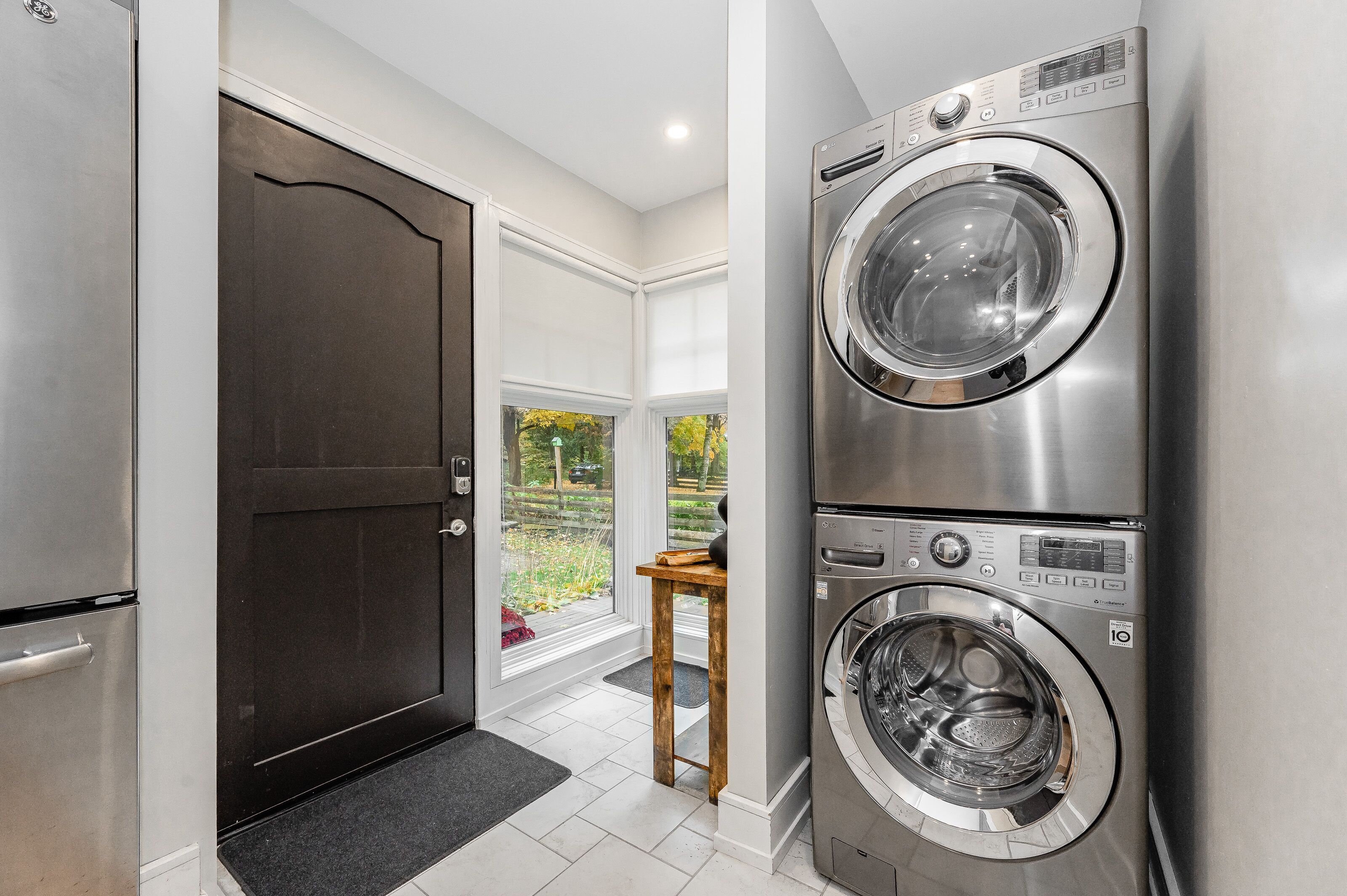 A laundry room with a double stainless steel washer and dryer, a dark door, and a view of a garden through a large window.