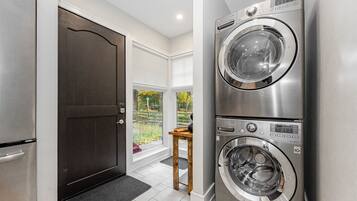 A laundry room with a double stainless steel washer and dryer, a dark door, and a view of a garden through a large window.