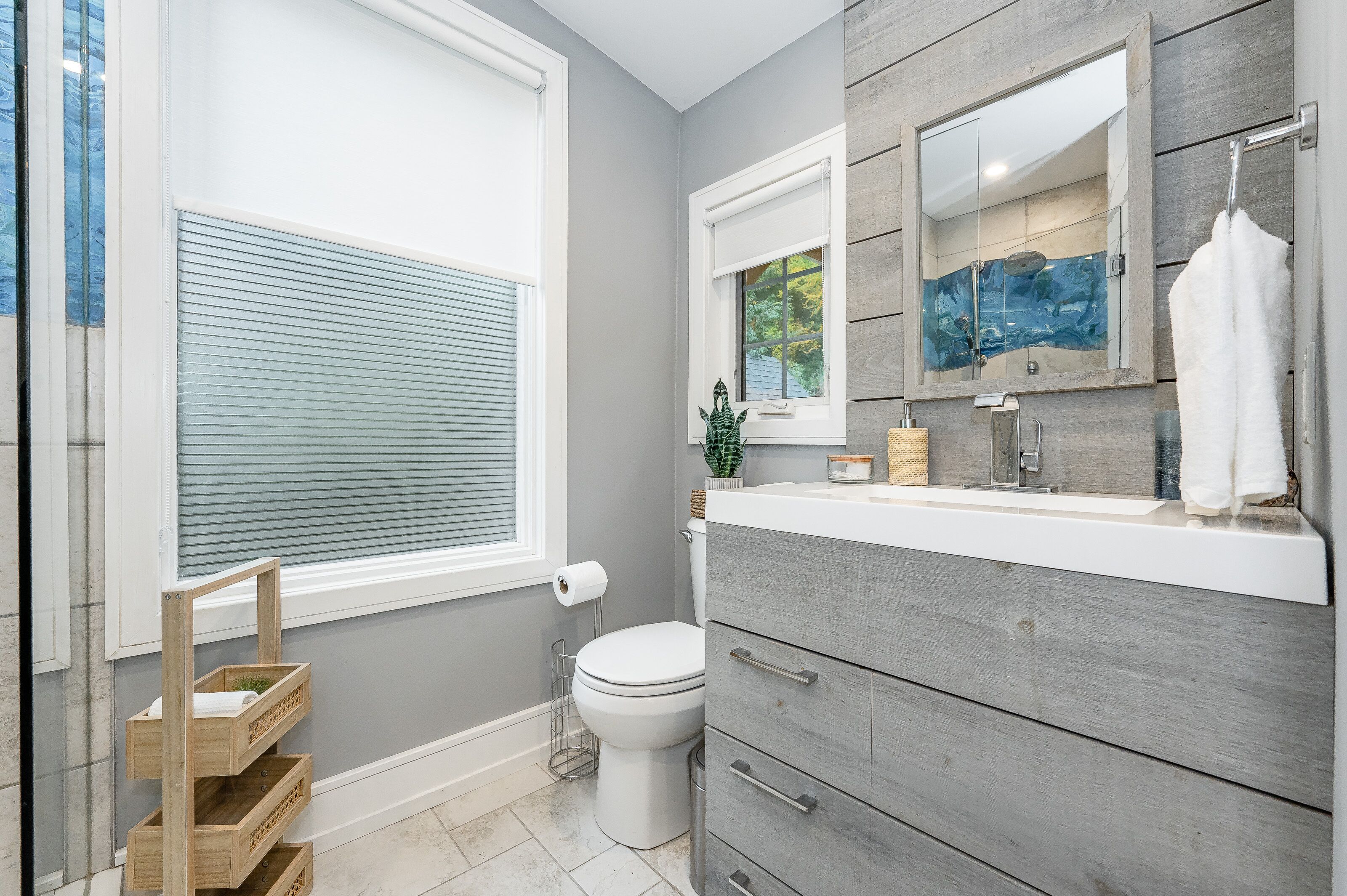 Une salle de bain équipée de toilettes, d’un lavabo et d’un meuble-lavabo. On y trouve une fenêtre avec des stores, un miroir et une serviette accrochée au mur.