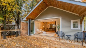Une terrasse en bois parsemée de feuilles d’automne, une porte vitrée donnant sur une pièce avec un canapé et des fauteuils, et une petite table.