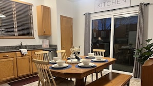 A kitchen with wooden cabinets, a dining table set with bowls and plates, a bench, and a sliding glass door leading to a dining area. - Spacious 6-BDR Near USAFA with Mountain Views *Pet Friendly* (Colorado Springs)