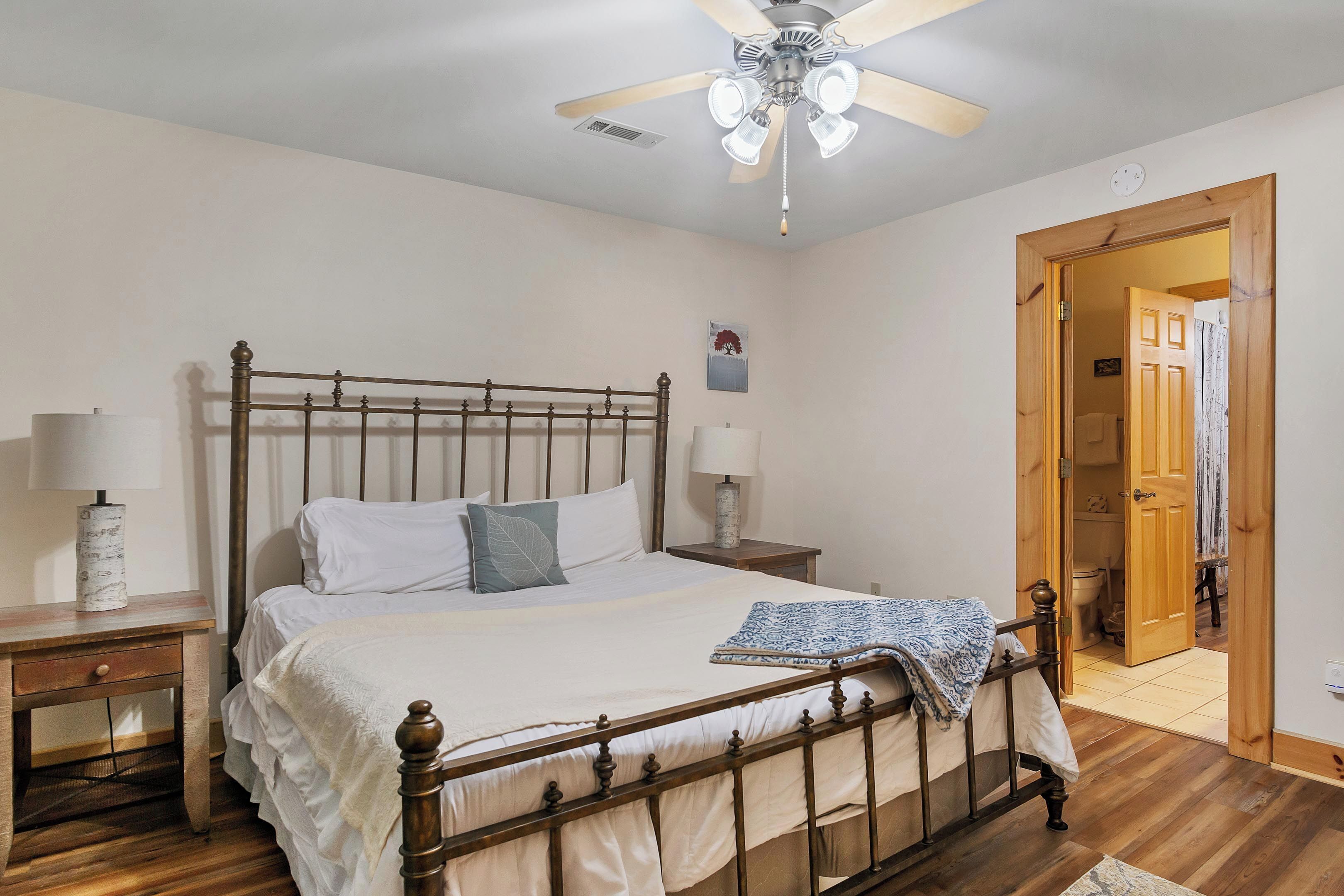 A bedroom with a wooden bed, two bedside tables with lamps, a ceiling fan, and a door leading to another room.