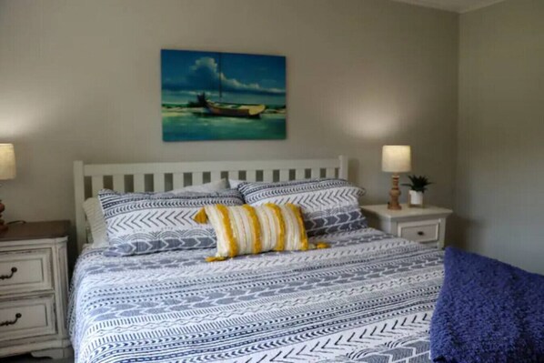 Un dormitorio con cama blanca, ropa de cama estampada, un cuadro de un barco en la pared y dos lámparas de noche.