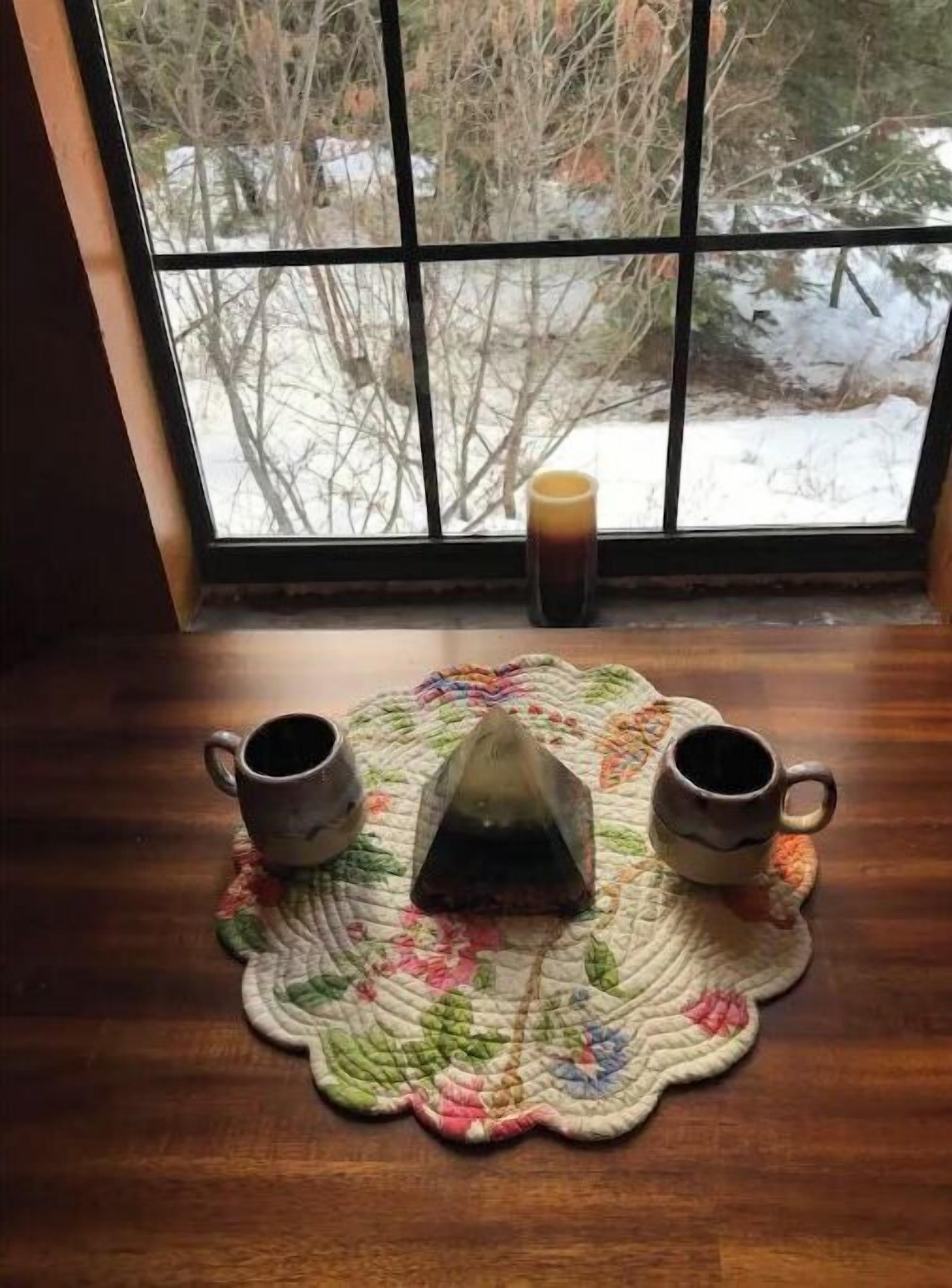 A window with a view of a snowy landscape, a wooden table with a placemat, and two ceramic mugs.