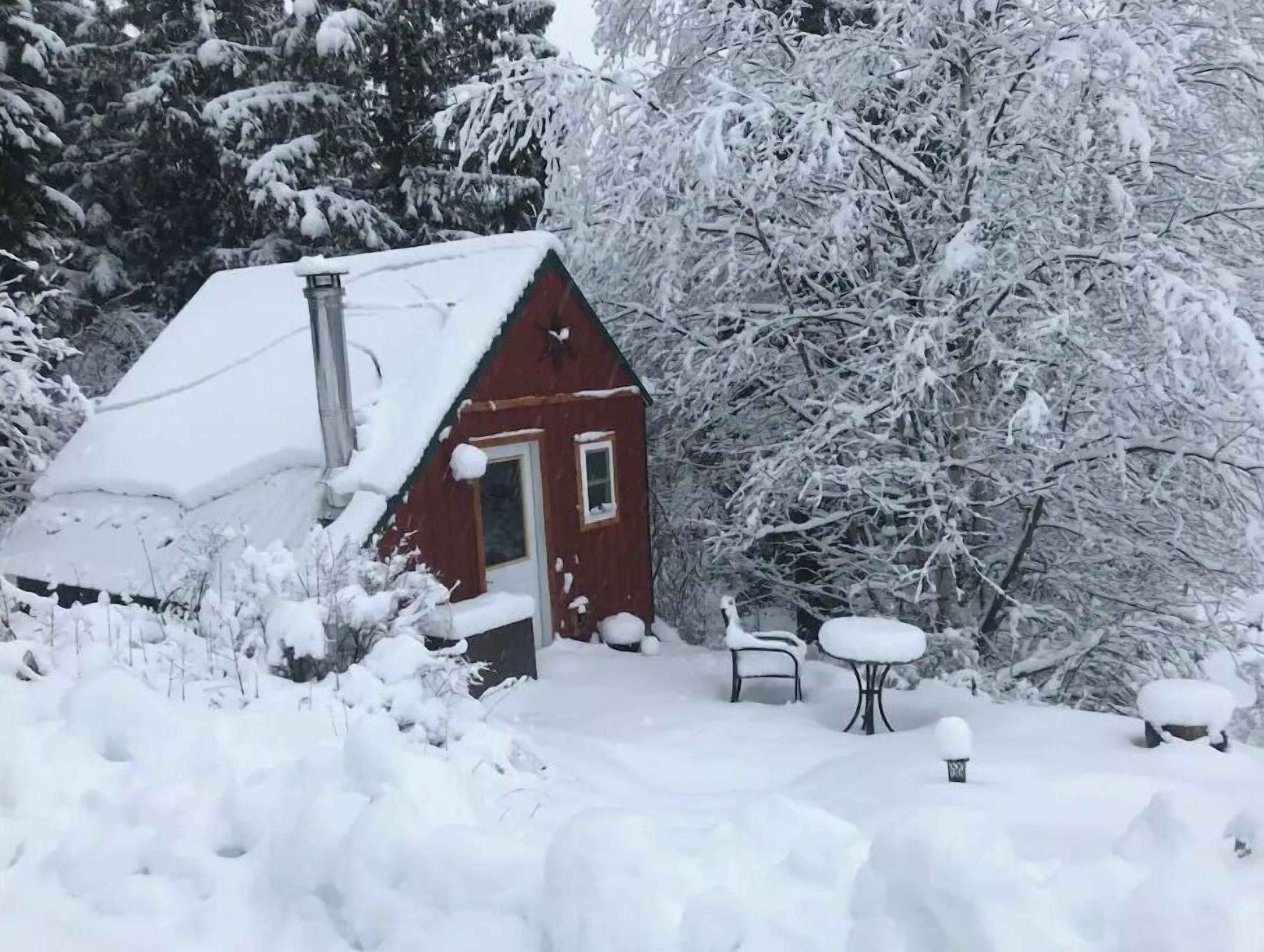 En liten, snötäckt stuga med röda väggar, en skorsten och en dörr. Utanför finns det två metallbord och bänkar, omgivna av snö.