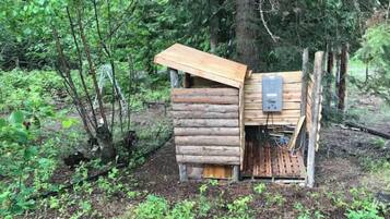 A rustic wooden cabin with a slanted roof, surrounded by dense forest.