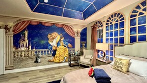 A room with a large mural of a fairy tale scene, a bed with a 'Be Our Guest' pillow, a night sky ceiling, and a view of a castle through the windows.