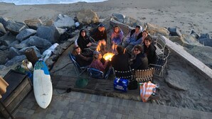Eine Gruppe von Menschen sitzt am Strand bei einem Lagerfeuer, in der Nähe liegen Surfbretter und eine Kiste Bud Light.