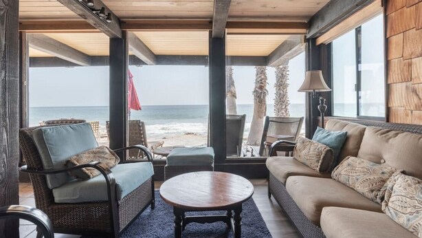 Ein gemütliches Zimmer mit Blick auf den Strand, ausgestattet mit Korbmöbeln, einem runden Couchtisch und einer Couch mit gemusterten Kissen.