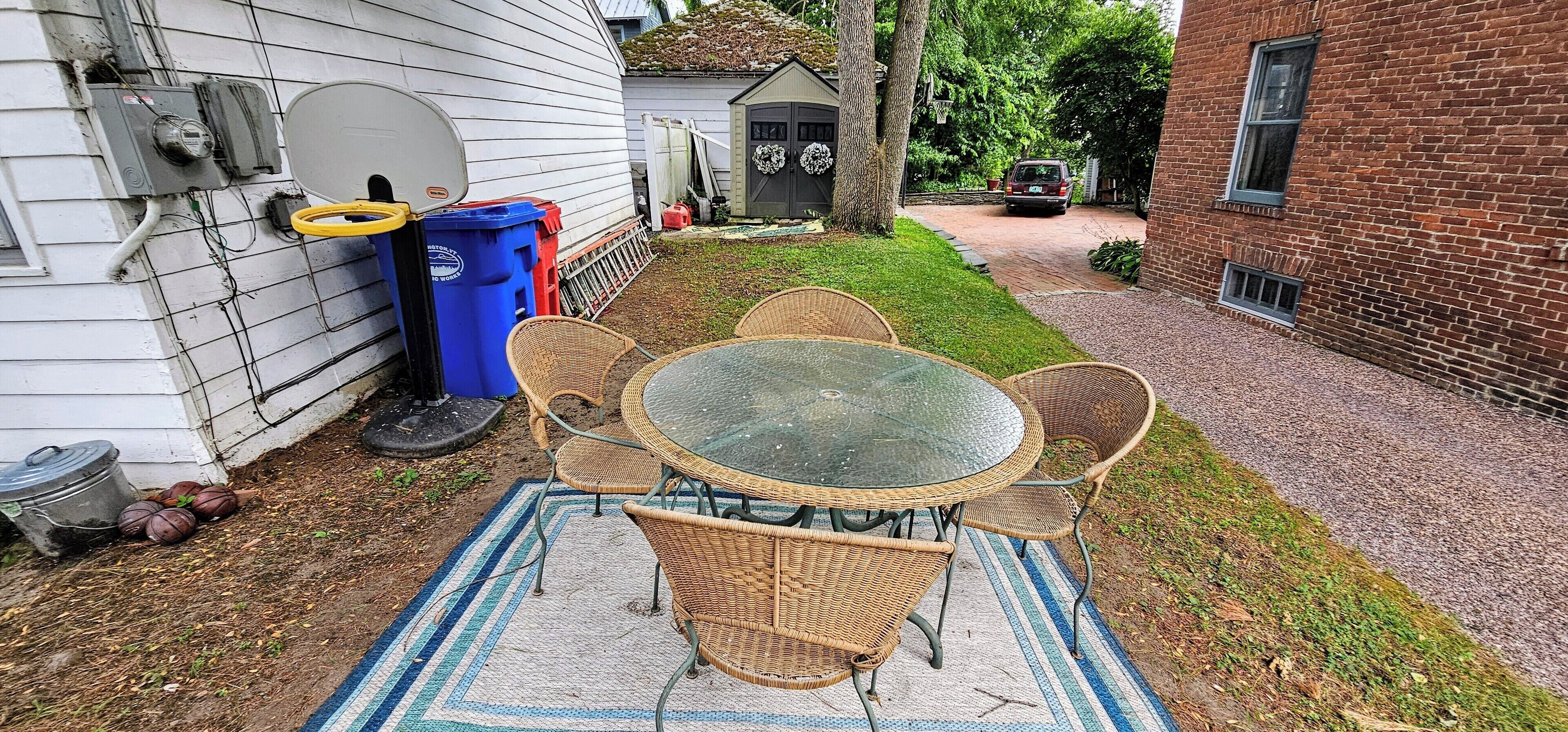 A patio with a glass-top table and wicker chairs, a blue and white striped rug, and a brick building in the background.