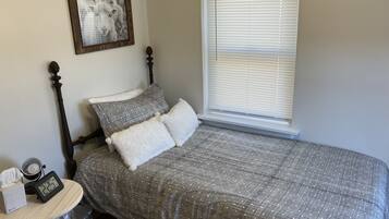 Un lit bien fait, recouvert d’une couverture à carreaux gris et blancs, avec deux oreillers, une table de chevet sur laquelle se trouvent une lampe et une horloge, et un tableau encadré accroché au mur.