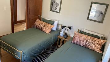 Una habitación con dos camas individuales, cada una con colcha verde y almohadas estampadas. Se ve una puerta de madera, una mesita de noche con lámpara, y cuadros enmarcados en la pared.