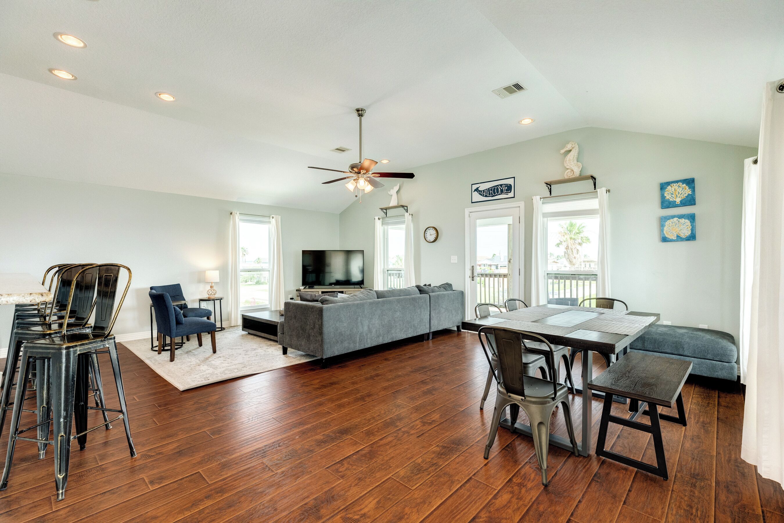 A spacious living room with hardwood flooring, a grey sofa, a dining table with chairs, and a ceiling fan.
