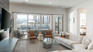 Un salon moderne doté d’un grand téléviseur, de sièges confortables, d’une table basse en verre et offrant une vue sur une piscine et des montagnes.