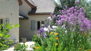 Une maison avec une façade en pierre, un toit en tuiles et un jardin où poussent différentes sortes de fleurs.