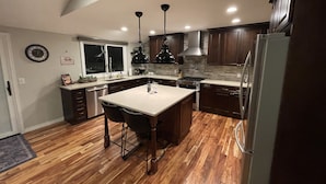 A modern kitchen with wooden flooring, a central island, dark wooden cabinets, and stainless steel appliances. - Willow Creek Oasis with Amazing Outdoor Kitchen (Centennial)