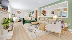 Ein modernes Wohnzimmer mit Kamin, einem grünen Sofa, weißen Sesseln und einem durch eine offene Tür einsehbaren Küchenbereich.