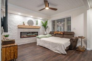 Un dormitorio moderno con chimenea, ventilador de techo, cama, televisor y ventana con persianas.