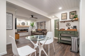 Una sala de estar moderna con una mesa de centro de vidrio, sillas blancas, chimenea, ventilador de techo y estación de café.