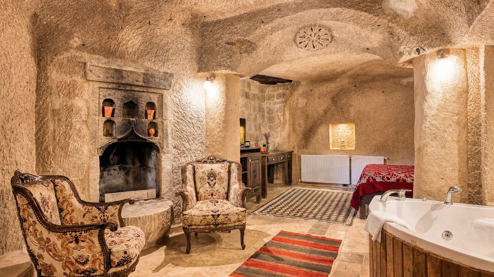 A room with a stone fireplace, two armchairs, a wooden cabinet, a red rug, and a bathtub.