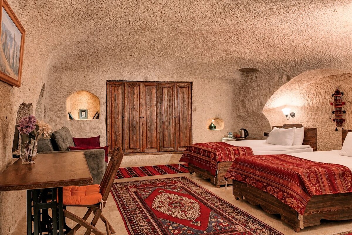 A room with a rustic stone wall, a wooden table with a chair, a bed with red bedding, and a small rug.
