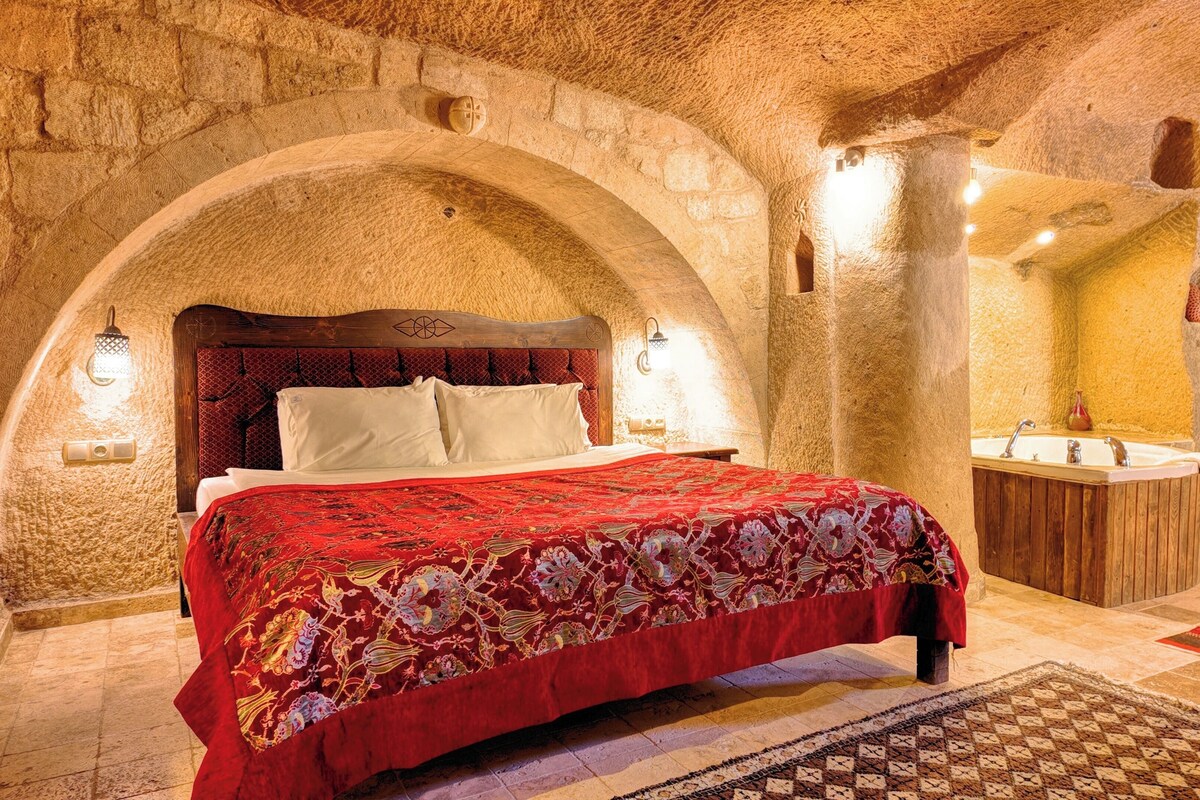 A room with a stone wall, a wooden bed with a red bedspread, a wooden nightstand, a wooden cabinet, and a small wooden table with a red and brown patterned rug.