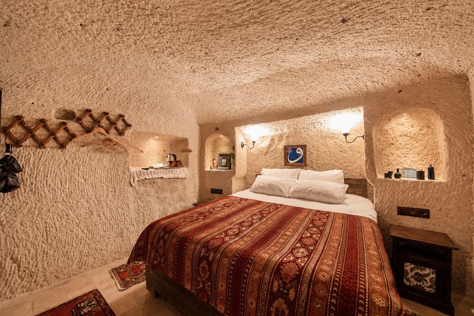 A room with a textured stone wall, a bed with a patterned bedspread, and a small shelf with decorative items.