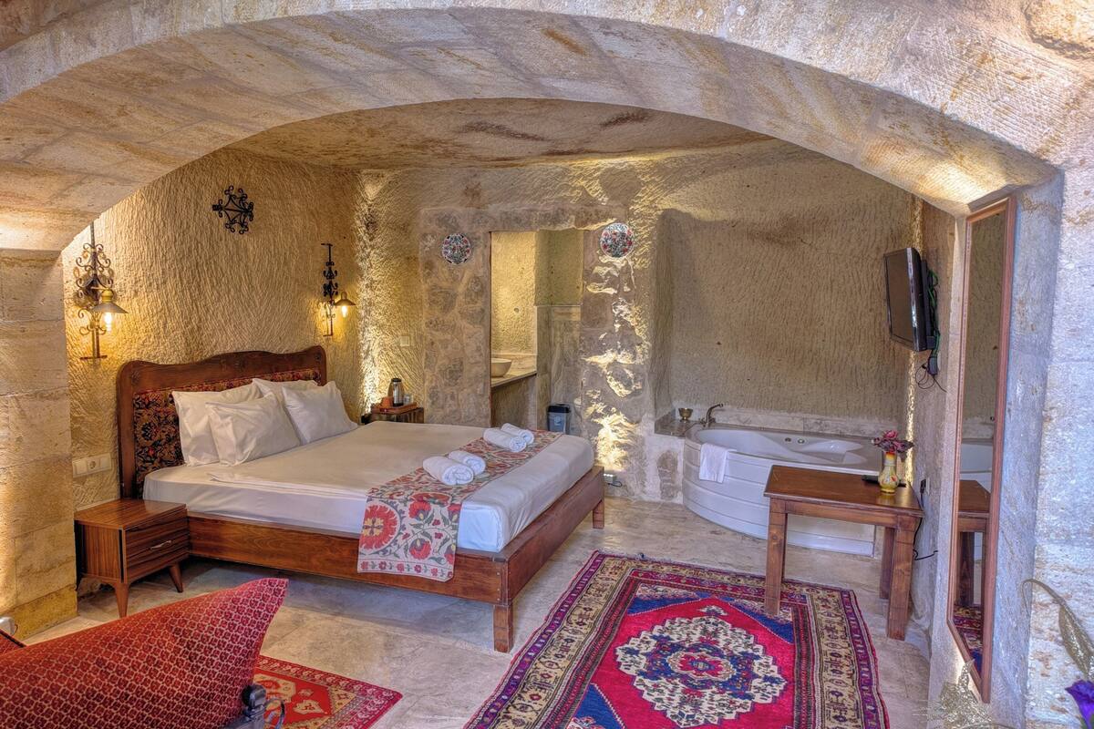 Quarto com paredes de pedra, cama de madeira, área de estar com sofá vermelho estampado, tapete grande e banheira.