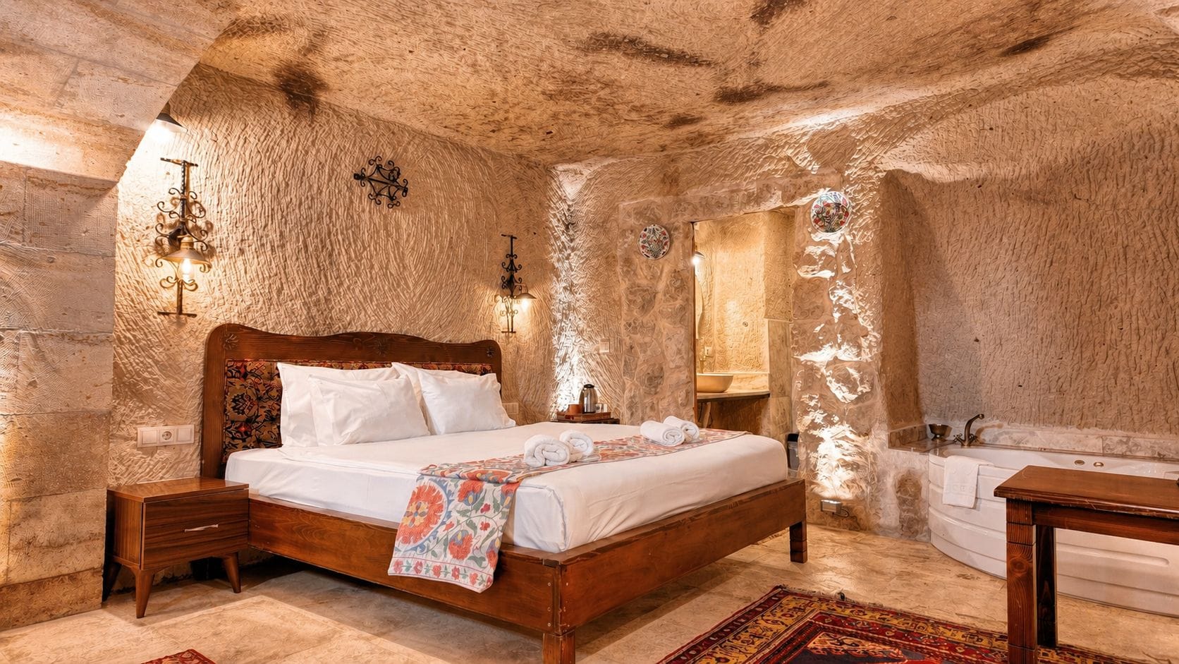 Een slaapkamer met een houten bed, nachtkastjes, een badkamer met een badkuip en een houten bankje.