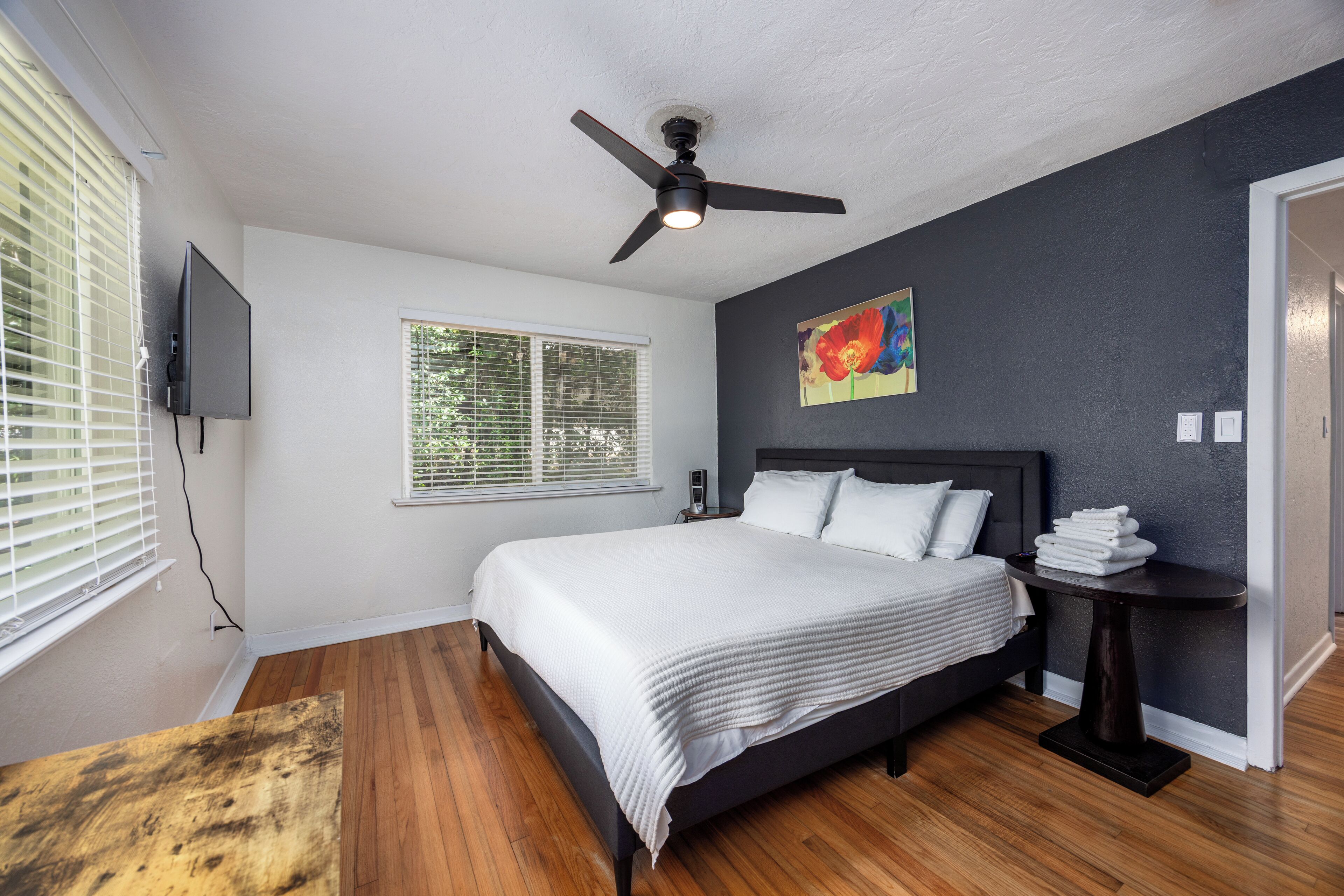 A bedroom with a bed, a ceiling fan, a window with blinds, a painting, and a nightstand with a lamp.