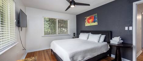 A bedroom with a bed, a ceiling fan, a window with blinds, a painting, and a nightstand with a lamp.