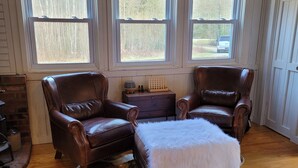 A room with two leather armchairs, a white fur ottoman, a wooden coffee table, and a chandelier. There are three windows with views of trees and a vehicle outside. - White mountain getaway (Carroll)