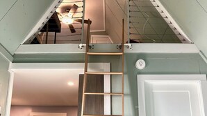 A loft bedroom with a wooden ladder leading to a mezzanine, a white door, and a kitchen area visible in the background. - Relaxing Scenic 1 Bedroom Retreat With Private Hot Tub (Findlay)