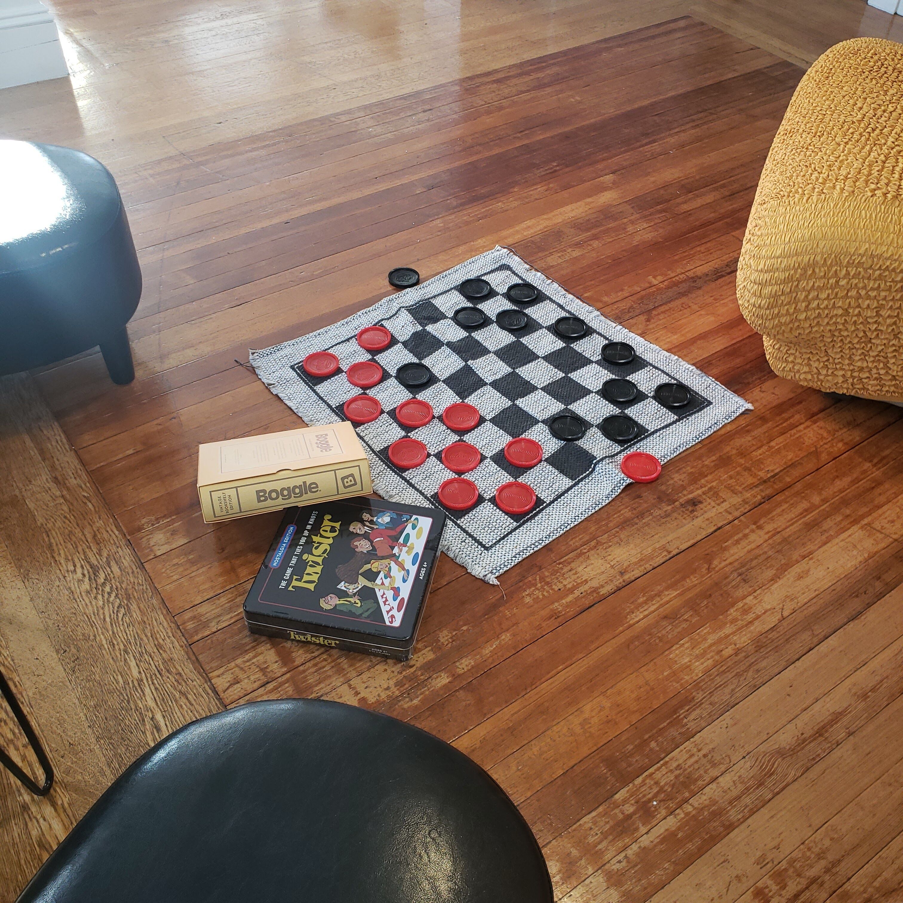 A checkered game board with red and black pieces, a board game box labeled "Boggle," and a "Twister" game box on a wooden floor.