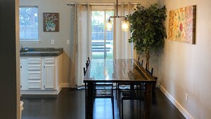 Un coin repas avec une table et des chaises en bois, un luminaire suspendu, une plante en pot et, au fond, une cuisine.