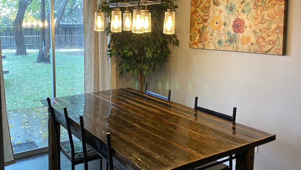 Un espace repas comprenant une grande table en bois, six chaises, un luminaire suspendu et une grande fenêtre donnant sur des arbres et une clôture.