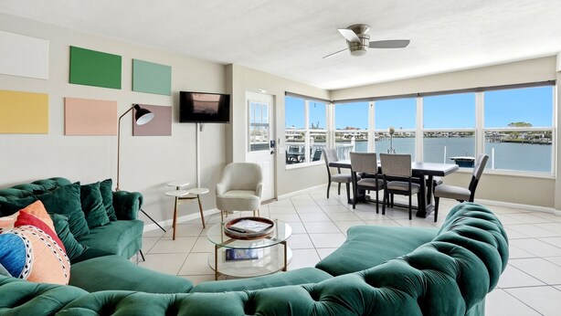 A modern living room with a green sectional sofa, a glass coffee table, a flat-screen TV, and a dining area with a view of the water.