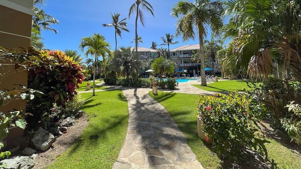 A pathway leading to a pool area surrounded by palm trees and a building with a tiled roof. - Nice 2 bedroom Apt. in Ocean Dream (Cabarete)