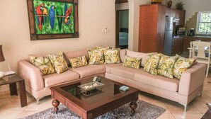 A living room with a sectional sofa, a glass coffee table, and a painting of parrots. - Nice 2 bedroom Apt. in Ocean Dream (Cabarete)
