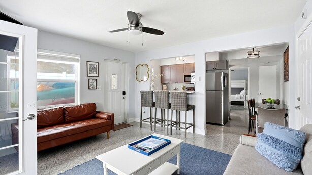 A modern living room with a brown leather sofa, a white coffee table, and a grey sofa with blue cushions. There is a kitchen area with bar stools and a refrigerator. - Waterfront condo overlooking the gorgeous intracoastal, Perfect for large family/group...sleeps up to 12 with 2 kitchens!  Well behaved dogs welcomed.  Bring your boat! (Indian Rocks Beach)
