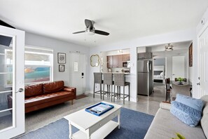 Una sala de estar moderna con un sofá de cuero marrón, una mesa de centro blanca y un sofá gris con cojines azules. Hay una zona de cocina con taburetes altos y un refrigerador.