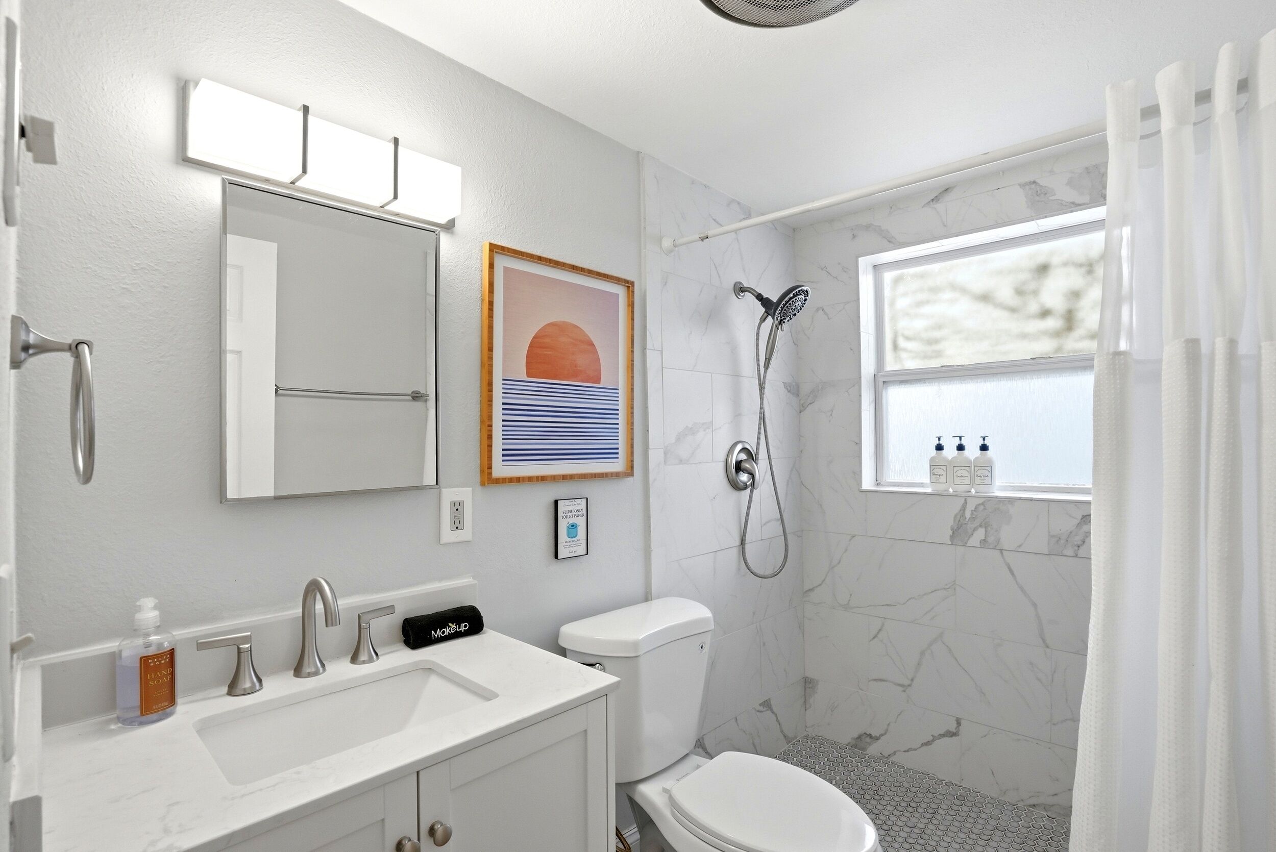 Une salle de bain avec un lavabo blanc, des toilettes, une douche, un tableau encadré et une fenêtre sur le rebord de laquelle sont posées des bouteilles.