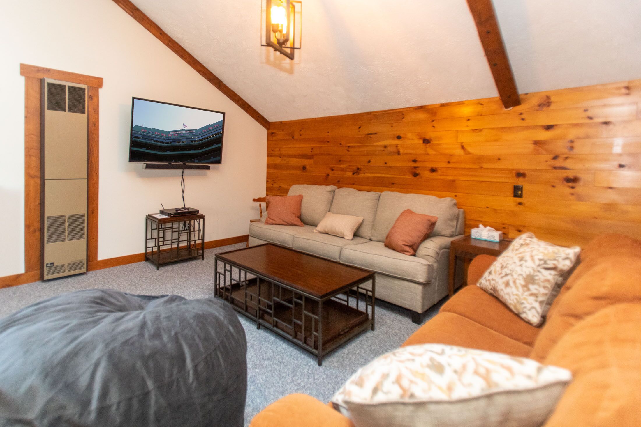 Een gezellige woonkamer met een houten plafond, een flatscreen televisie aan de muur, een beige bank met kussens, een houten salontafel en een grote relaxfauteuil.